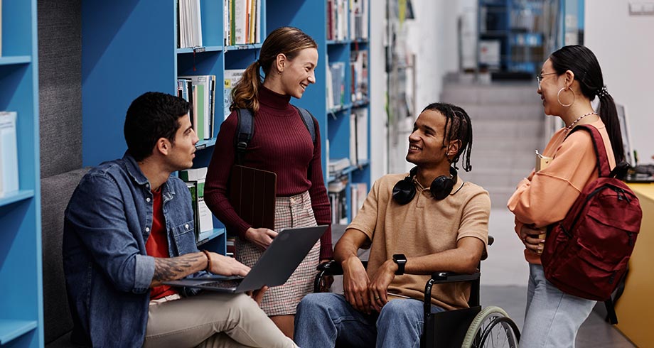 Diverse group of students with young man in wheelchair chatting cheerfully in college library